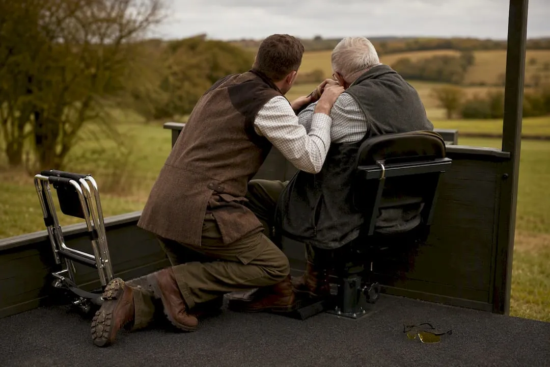 A coach kneels beside an older shooter seated in a swivel chair on a covered shooting stand overlooking fields, with a folded walking frame beside them