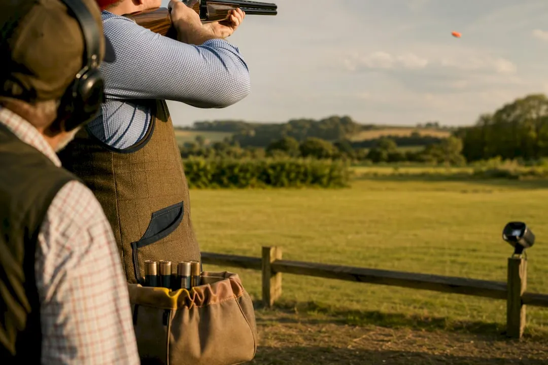 A shooter in a tweed vest mounts an over-and-under shotgun as an orange clay flies over a sunlit field, with an onlooker in ear defenders in the foreground