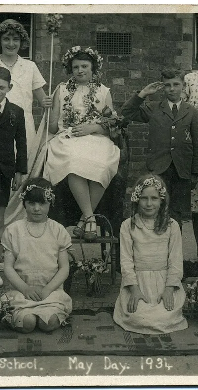 1934 postcard of Chiseldon School, near Swindon, Wiltshire, England, showing a group of school children dressed for May Day celebrations. It is one of a set of seven postcards from the 1930s, showing children from the same school on May Day.  There is a May Queen; also Brownies, Cubs, Scouts and Girl Guides.
The photographer was Fred C. Palmer of Tower Studio, Herne Bay, Kent ca.1905-1920, and of 6 Cromwell Street, Swindon ca.1920-1936. He is believed to have died 1936-1939. 
This print has darkened with age, but it would be inappropriate to adjust the brightness because detail, e.g. on white dresses, would be lost.

Border
The remaining border of this image is important for researchers of this photographer. Some photographers trimmed their images more than others, and Palmer has a reputation for producing smaller postcards than other early 20th century UK photographers. He took his own photos, developed them in-house onto postcard-backed photographic paper and trimmed them himself. It is worth adding that during hand-developing the border is actively masked with equipment which both crops the picture and causes the white frame or border to appear on the paper. This frame is part of the design and is one of the reasons why the quality of Palmer's work is so interesting, and why there is an article and category for him on English Wiki. Researchers need to see exactly where the edge of the postcard is. Thank you for taking the time to read this.