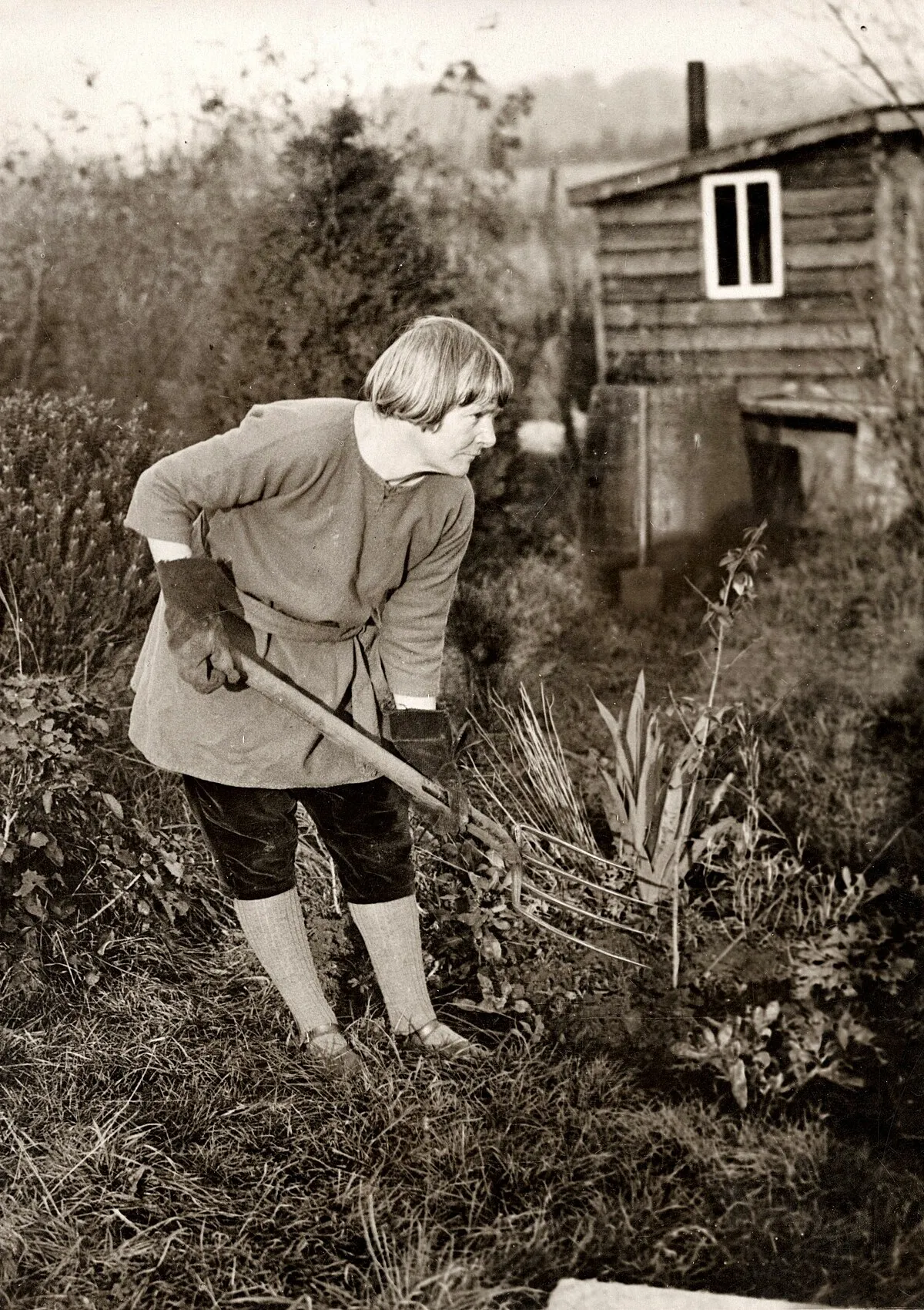 Omschrijving: Natuurmensen 1924. Kolonie van 'primitieven' in Engeland. man is in de tuin aan het werk voor zijn houten huisje. Een regenton vangt het water op.