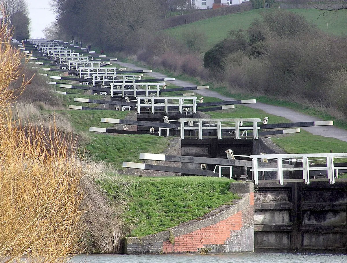 The Caen Hill locks, on the Kennet and Avon Canal, Devizes, Wiltshire, England, in early Spring. The sixteen locks of Caen Hill (seen in this picture), and a further 13 locks, stretch over only 2 miles of the canal. The large side ponds are not seen here. The white "fences" are bridges across each lock.
The building of these locks in 1810 marked completion of the whole 57 mile distance from Newbury to Bath. 
Photographed by Adrian Pingstone in March 2007 and placed in the public domain.
Other versions:
