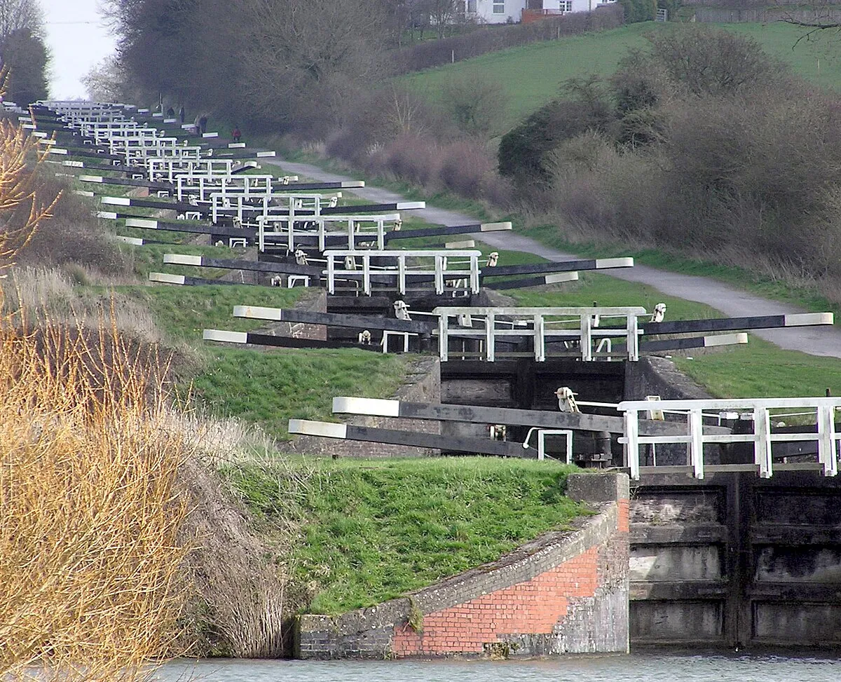 The Caen Hill locks, on the Kennet and Avon Canal, Devizes, Wiltshire, England, in early Spring. The sixteen locks of Caen Hill (seen in this picture), and a further 13 locks, stretch over only 2 miles of the canal. The large side ponds are not seen here. The white "fences" are bridges across each lock.
The building of these locks in 1810 marked completion of the whole 57 mile distance from Newbury to Bath. 
Photographed by Adrian Pingstone in March 2007 and placed in the public domain.
Other versions: