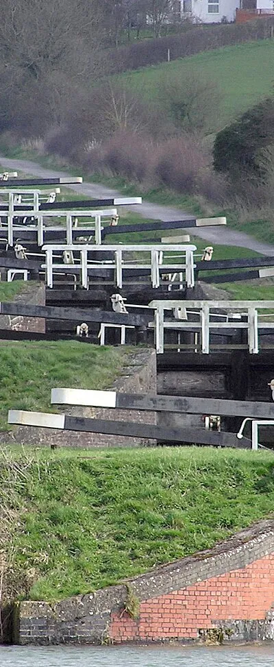 The Caen Hill locks, on the Kennet and Avon Canal, Devizes, Wiltshire, England, in early Spring. The sixteen locks of Caen Hill (seen in this picture), and a further 13 locks, stretch over only 2 miles of the canal. The large side ponds are not seen here. The white "fences" are bridges across each lock.
The building of these locks in 1810 marked completion of the whole 57 mile distance from Newbury to Bath. 
Photographed by Adrian Pingstone in March 2007 and placed in the public domain.
Other versions: