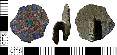 A copper-alloy Early Medieval hanging bowl mount or escutcheon probably dating to the c. 7th century AD. The mount consists of a discoidal mount body, with a broken hanging terminal. The terminal is decorated with three circles two of which are sub-divided into recessed squared segments decorated with milifiori glass composed of blue with white dots delineated by red. Within the very centre of the mount, a recessed circle has been inlaid with green glass or enamel. The outer circumference of the mount disc itself has also been tinned or silvered.

Millefiori is very unusual on hanging-bowl mounts; Bruce-Mitford lists 11 examples of hanging-bowls with millefiori (2005, 67). There are similar star-in-circle designs from several, such as the St Paul in the Bail bowl, the exterior base escutcheon on the Manton Warren bowl, and on the Sutton Hoo bowl 1 (Bruce-Mitford 2005, col pl. 4); the same design can also be found in an enamelled version on the Baginton bowl (Bruce-Mitford 2005, no. 93).
The length is 29mm, the width is 25mm thickness 10mm. Photos from finder.