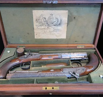 Pair of cased Joseph Manton percussion duelling pistols (1820s-1840s) two identical percussion pistols; .50 inch calibre; single octagonal iron barrel; hooked breech; captive ramrod; chequered walnut one piece grip with iron butt plate; silver oval escutcheon, unmarked, on wrist; engraved on trigger guard and on butt; action crisp accessories- case- rectangular wooden case; lid has concentric brass circles; lined with green baize with paper label on interior lid with lion, unicorn, and coat of arms; case has seven compartments, two with lids; partitions are loose bullet mould- steel; numbered 27 on handle oil bottle- steel; cylindrical powder flask- copper and brass; adjustable powder measure at spout- 3-4, 5-8, 1-2 brass cap mallet shaped ramrod- with brass fittings; with attached worm and additional patch-cleaning jag; leather inserts at each end of mallet head markings- on lock plate- JOSEPH MANTON - LONDON printed paper label inside case- Joseph Manton - GUN MAKER - Davies Street Berkeley Square - LONDON - Patentee for the Elevation of Double Guns, Hammers of Locks, - Inverted Breeching, and Gravitating Stops manufacturer Joseph Manton, 1795-1835