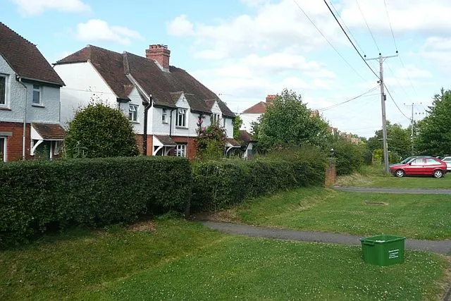 1950s houses in Leckhampstead This long row of houses (about 30 in groups of three) is actually at an angle to the road. So the verge where the red car is parked gets wider as one goes east until at the far end a separate road has been created.