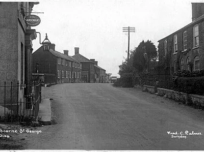 Photograph no. 0104 by Frederick Christian Palmer of Swindon (d.1941), looking south-east on Marlborough Road, in Ogbourne St George, Wiltshire, England. The probable public house or hotel on the left exists today as a pub, restaurant and guest house, although the railings, door lamp and Sandemans sign have been removed. The houses on the right also exist, although those farther along the road at the left have been demolished and redeveloped.