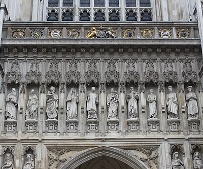 Westminster Abbey (The Collegiate Church of St Peter). Ten 20th-century martyrs from across the world are depicted in statues erected in 1995 above the Great West Door of Westminster Abbey. Nine heraldic shields, from reign of King George II. The abbey's two western towers were built between 1722 and 1745 by Nicholas Hawksmoor, constructed from Portland stone to an early example of a Gothic Revival design. In 1723, on the death of Wren, he became architect to Westminster Abbey, the west gable (1735) and towers (1734-1745) of which are his. Heraldry, left to right:
1: Order of the Bath. The Lady Chapel is associated with the Order of the Bath
2: Nassau? The coat of arms of the House of Nassau is prominently associated with King William III (William of Orange), who was crowned King of England, Scotland, and Ireland in Westminster Abbey in 1689
3: Kingdom of Middlesex (and of Essex)
4: Royal Arms of England 1406-1603 (possibly intended for King Henry VII (1485-1509) who built the Lady Chapel)
5: (centre) Royal arms of King George II (1727-1760) (Royal arms of Great Britain (1714-1801)). Identified as King George II because the arms of  w:Joseph Wilcocks (1673-1756)  Dean of Westminster 1731-56 (presumably when this element was built), also appear (see 6), and he served as Dean entirely during the reign of King George II
6: Coat of arms of Westminster Abbey impaling Ermine, a chief chequy azure and or, arms of w:Joseph Wilcocks (1673-1756)  Dean of Westminster 1731-56 (presumably when this element was built), Bishop of Gloucester, Bishop of Rochester (Burke, Sir Bernard, The General Armory, London, 1884, p.1110 "Wilcox alias Nowers, Leicestershire, 1732", but with chief chequy or and sable), another or and gules)
7: Coat of arms of the City of Westminster originally granted 1 October 1601 to the Borough of Westminster "Quarterly: 1st and 4th, Azure a Portcullis with chains pendant Or on a Chief of the last between two united Roses of York and Lancaster a Pale charged with the Arms of King Edward the Confessor (i.e. the old arms of the City of Westminster originally granted 1 October 1601 to Borough of Westminster, regranted by royal warrant dated 6 November 1901 to Metropolitan borough. Source: A.C. Fox-Davies, The Book of Public Arms (T. C. &amp; E. C. Jack, London, 1915. See 25px|Westminster arms)); Now borne by the Duke of Westminster
8: Attributed arms of the Kingdom of Kent (or: Duchy of Saxony (as in the Lesser Arms of Hanover as borne by Kings of the House of Hanover (as at centre (5)))
9: A rose and thistle conjoined in a single stem (presumably signifying the union of the Kingdoms of England and Scotland)