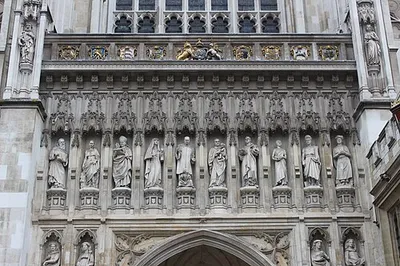Westminster Abbey (The Collegiate Church of St Peter). Ten 20th-century martyrs from across the world are depicted in statues erected in 1995 above the Great West Door of Westminster Abbey. Nine heraldic shields, from reign of King George II. The abbey's two western towers were built between 1722 and 1745 by Nicholas Hawksmoor, constructed from Portland stone to an early example of a Gothic Revival design. In 1723, on the death of Wren, he became architect to Westminster Abbey, the west gable (1735) and towers (1734-1745) of which are his. Heraldry, left to right:
1: Order of the Bath. The Lady Chapel is associated with the Order of the Bath
2: Nassau? The coat of arms of the House of Nassau is prominently associated with King William III (William of Orange), who was crowned King of England, Scotland, and Ireland in Westminster Abbey in 1689
3: Kingdom of Middlesex (and of Essex)
4: Royal Arms of England 1406-1603 (possibly intended for King Henry VII (1485-1509) who built the Lady Chapel)
5: (centre) Royal arms of King George II (1727-1760) (Royal arms of Great Britain (1714-1801)). Identified as King George II because the arms of w:Joseph Wilcocks (1673-1756) Dean of Westminster 1731-56 (presumably when this element was built), also appear (see 6), and he served as Dean entirely during the reign of King George II
6: Coat of arms of Westminster Abbey impaling Ermine, a chief chequy azure and or, arms of w:Joseph Wilcocks (1673-1756) Dean of Westminster 1731-56 (presumably when this element was built), Bishop of Gloucester, Bishop of Rochester (Burke, Sir Bernard, The General Armory, London, 1884, p.1110 "Wilcox alias Nowers, Leicestershire, 1732", but with chief chequy or and sable), another or and gules)
7: Coat of arms of the City of Westminster originally granted 1 October 1601 to the Borough of Westminster "Quarterly: 1st and 4th, Azure a Portcullis with chains pendant Or on a Chief of the last between two united Roses of York and Lancaster a Pale charged with the Arms of King Edward the Confessor (i.e. the old arms of the City of Westminster originally granted 1 October 1601 to Borough of Westminster, regranted by royal warrant dated 6 November 1901 to Metropolitan borough. Source: A.C. Fox-Davies, The Book of Public Arms (T. C. & E. C. Jack, London, 1915. See 25px|Westminster arms)); Now borne by the Duke of Westminster
8: Attributed arms of the Kingdom of Kent (or: Duchy of Saxony (as in the Lesser Arms of Hanover as borne by Kings of the House of Hanover (as at centre (5)))
9: A rose and thistle conjoined in a single stem (presumably signifying the union of the Kingdoms of England and Scotland)
