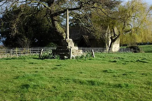 Base and shaft of a Medieval stone cross at Wick, Worcestershire, with St Mary's parish church visible through the trees